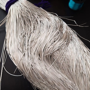 Könnte beinhalten: Nahaufnahme eines Bündels silberfarbenen Drahtes, zusammengebunden mit einem dunkelvioletten Band. Der feine Draht hat einen leichten Glanz. Im Hintergrund sind mehrere Spulen farbigen Garns zu sehen, was auf ein Bastel- oder Schmuckthema hindeutet.