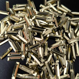 Peut inclure: Un ensemble de petits strass rectangulaires dorés avec un éclat métallique. Chaque strass a un dos plat et un dessus facetté, créant un effet scintillant. Les strass sont dispersés sur une surface sombre.
