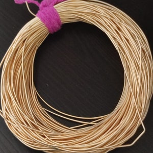 Könnte beinhalten: Eine Spule aus goldfarbenem Draht, eng gewickelt und mit einem rosa, flauschigen Band gesichert. Der Draht ist glatt und reflektierend, was auf ein metallisches Material hindeutet. Das Bild ist eine Nahaufnahme, die die Textur und Farbe des Drahtes hervorhebt.