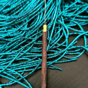 Può includere: Uno strumento per aghi in legno con una punta in metallo, appoggiato su un mucchio di filo blu turchese.