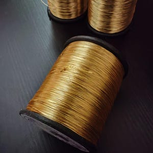 Puede incluir: Tres carretes de hilo dorado, enrollados apretadamente en bobinas de plástico negro. El hilo es brillante y metálico.