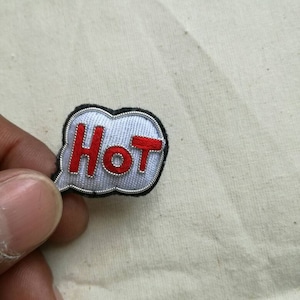Peut inclure: Écusson brodé avec le mot "HOT" en rouge à l'intérieur d'une bulle de dialogue blanche bordée de noir. Le patch est tenu par une main sur un fond de tissu neutre.