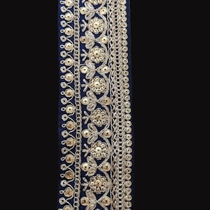 May include: Gold and silver embroidered trim on a dark blue fabric background. The trim features a repeating pattern of floral motifs and small beads.