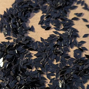 Peut inclure: Gros plan sur une collection de petits éléments noirs en forme de losange, dispersés sur une surface brun clair. Les pièces sont réfléchissantes, captant la lumière. L'arrangement suggère un matériau décoratif ou artisanal.