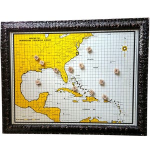 May include: A framed magnetic hurricane tracking chart with a yellow and white map of the southeastern United States and the Caribbean. Wooden letter tiles spell out "hurricane" on the chart. The frame is dark brown with an ornate design.