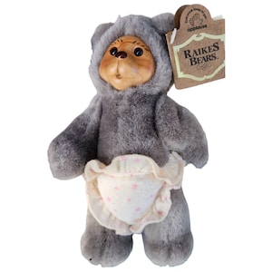 Może przedstawiać: Pluszowa zabawka Raikes Bears, przedstawiająca szarego misia w kapturze i pieluszce z różowymi gwiazdkami. Miś ma drewnianą twarz i metkę z napisem "Raikes Bears."
