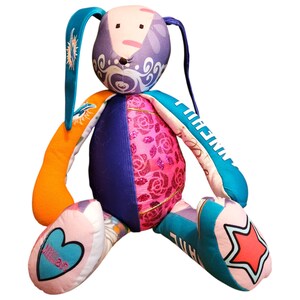 Può includere: Un coniglietto di peluche fatto a mano e colorato con un design patchwork. Il coniglietto presenta una testa viola con vortici bianchi, braccia arancioni e blu e un busto rosa con motivi floreali. I piedi hanno un design a cuore e stella. Il nome "Jillian" è su un piede.