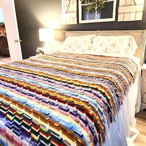 May include: A vibrant, hand-crocheted blanket with a zig-zag design. The blanket displays a spectrum of colours, including blue, purple, yellow, and red. It is placed on a bed with floral pillows and a white bed skirt.