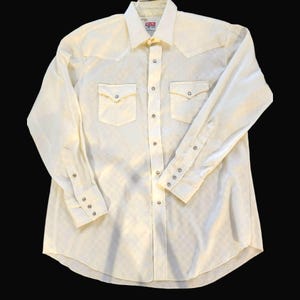 Puede incluir: Camisa crema de manga larga con estampado de cuadros. La camisa tiene dos bolsillos en el pecho, cierres de presión y un cuello clásico. Los puños también tienen cierres de presión. La camisa es de estilo occidental.