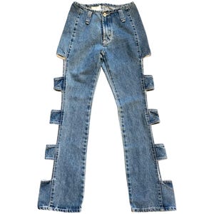 May include: Light blue denim jeans with unique cut-out details along the outer seams. The jeans have a classic five-pocket design, a button and zipper closure, and belt loops. The denim has a faded wash.