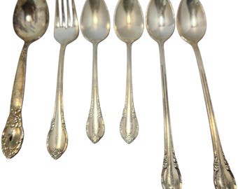 Op de afbeelding: Een verzameling vintage zilverwerk, waaronder lepels en een vork, gerangschikt tegen een witte achtergrond. De lepels hebben ovale bakken en sierlijke handvatten. De vork heeft vier tanden. Het zilverwerk lijkt van zilver of een zilverkleurig metaal te zijn.