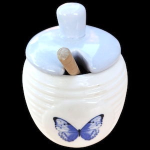 May include: A white ceramic honey pot with horizontal ribbed detailing and a blue butterfly graphic on the front. The pot includes a matching light blue lid with a central knob and a notched opening, containing a wooden honey dipper inside.