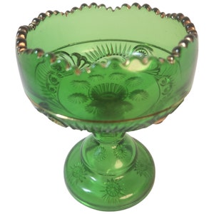 May include: A green glass pedestal bowl with a scalloped rim and gold trim. The bowl features embossed floral and geometric patterns. The base is round and also has embossed floral designs.