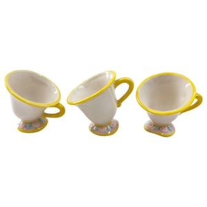Könnte beinhalten: Drei weiße Keramik-Teetassen mit gelben Griffen und Rändern. Der Boden jeder Tasse ist mit pastellfarbenen Blumendesigns verziert. Die Teetassen sind in verschiedenen Winkeln geneigt.