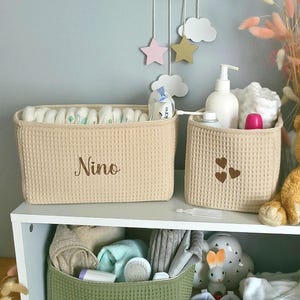 Peut inclure: Deux paniers de rangement en tissu beige à texture gaufrée, l'un rectangulaire avec le nom Nino brodé et l'autre carré avec trois petits cœurs marron, accompagnés d'un ours en peluche, d'articles de puériculture et d'un panier vert.