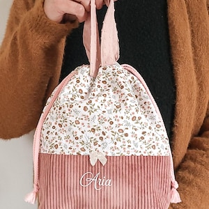 May include: A drawstring pouch featuring a dusty pink corduroy base and a white upper section with a small floral print. The bag includes pink rope handles, a small cream-colored bow, and the name Aria embroidered in white script on the front.