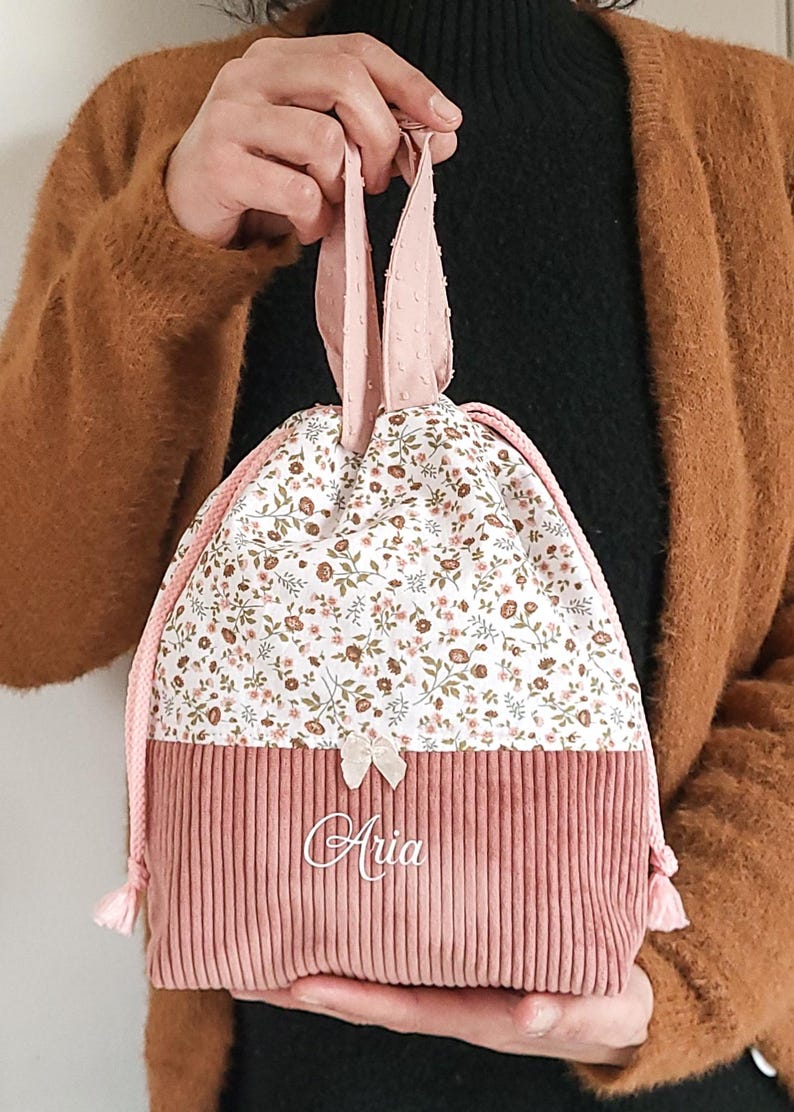 May include: A drawstring pouch featuring a dusty pink corduroy base and a white upper section with a small floral print. The bag includes pink rope handles, a small cream-colored bow, and the name Aria embroidered in white script on the front.