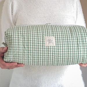 Può includere: Un beauty case rettangolare trapuntato con un motivo a quadretti verdi e bianchi. La borsa ha una chiusura a cerniera e una piccola etichetta con la scritta "Atelier D50". La borsa è tenuta da una persona.