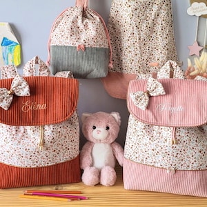 May include: Two corduroy backpacks with floral fabric panels, one rust-colored with the name Elena and one pink with the name Fanyette, each featuring a matching bow on the flap, alongside a drawstring bag and a plush pink teddy bear.