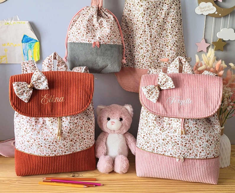 May include: Two corduroy backpacks with floral fabric panels, one rust-colored with the name Elena and one pink with the name Fanyette, each featuring a matching bow on the flap, alongside a drawstring bag and a plush pink teddy bear.