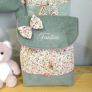 May include: A sage green corduroy backpack featuring a floral print fabric panel, a white bow with matching floral pattern, and the name Fantine embroidered in white script on the front flap. The bag includes pink drawstring cords and trim.