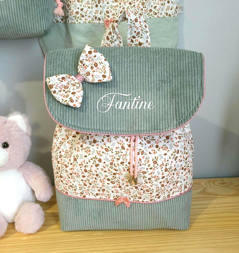 May include: A sage green corduroy backpack featuring a floral print fabric panel, a white bow with matching floral pattern, and the name Fantine embroidered in white script on the front flap. The bag includes pink drawstring cords and trim.