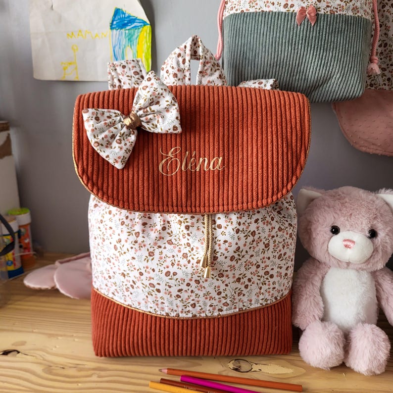 May include: A rust-colored corduroy backpack featuring a floral print panel, a matching floral bow on the flap, and the name Elena embroidered in gold script. The bag includes a drawstring closure with gold-toned metal tips and a top handle.