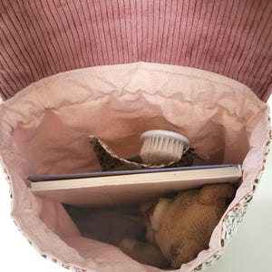 May include: A backpack with a dusty rose corduroy flap and a light pink fabric interior. The bag contains a white-bristled brush, a rectangular book with a light blue cover, and a small brown plush bear tucked inside the main compartment.