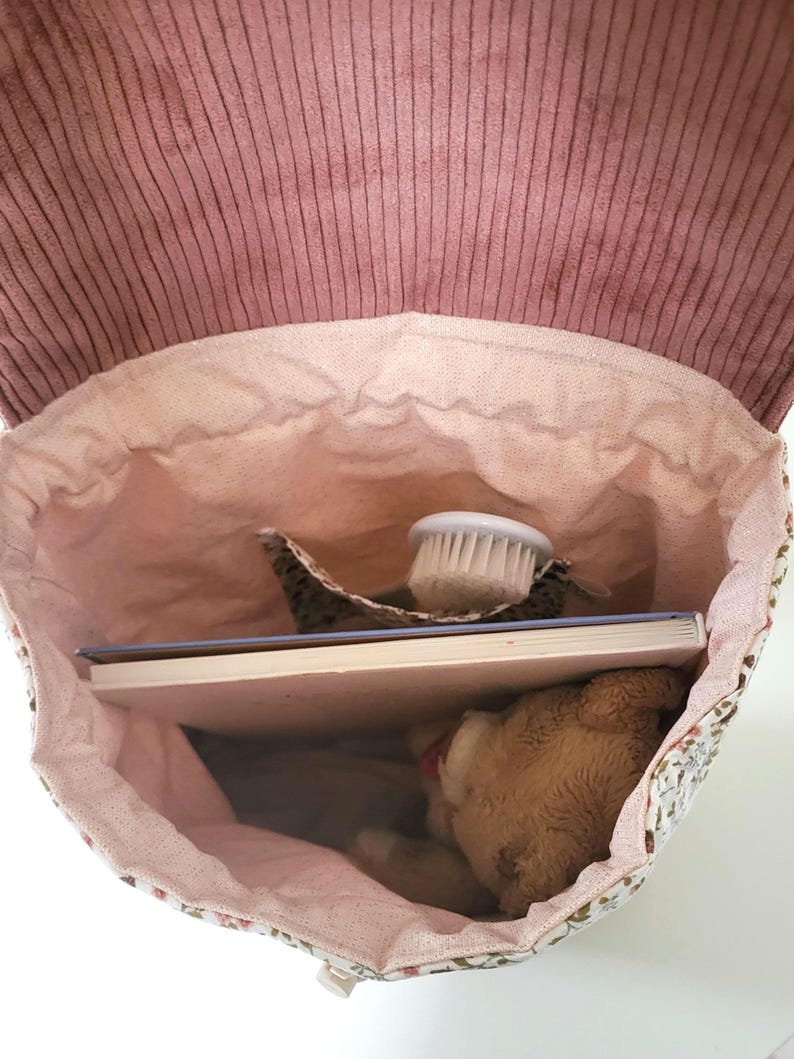 May include: A backpack with a dusty rose corduroy flap and a light pink fabric interior. The bag contains a white-bristled brush, a rectangular book with a light blue cover, and a small brown plush bear tucked inside the main compartment.