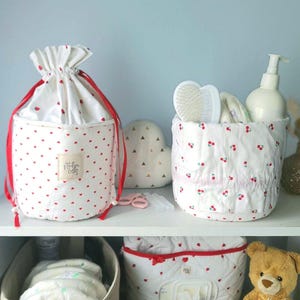 Peut inclure: Un sac à cordon en tissu blanc avec un motif de cœurs rouges et une étiquette carrée portant l'inscription "helen dse" à côté d'un panier en tissu blanc à motif de cerises rouges contenant une brosse et un flacon pompe blanc.
