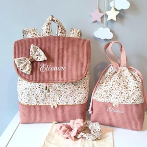 Kit maternelle fille avec un Ensemble sac à dos  en velours côtelé vieux rose personnalisé avec le prénom Eléanore en blanc avec des imprimés floraux et un noeud sur le rabat. Un sac à goûter personnalisé avec le prénom et Des chouchous assortis sont également inclus.
