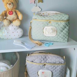 Può includere: Due pochette rettangolari trapuntate per salviette con bordo dorato e cinturino da polso. La pochette superiore è verde salvia con un dispenser bianco, quella inferiore è grigia con un dispenser bianco con logo floreale e le lettere DS.