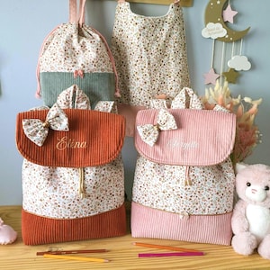 May include: Two corduroy backpacks with floral fabric panels, one in rust orange and one in light pink. Each features a matching floral bow on the flap, a drawstring closure, and embroidered names Elena and Feryelle. A matching drawstring bag and bib are visible in the background.