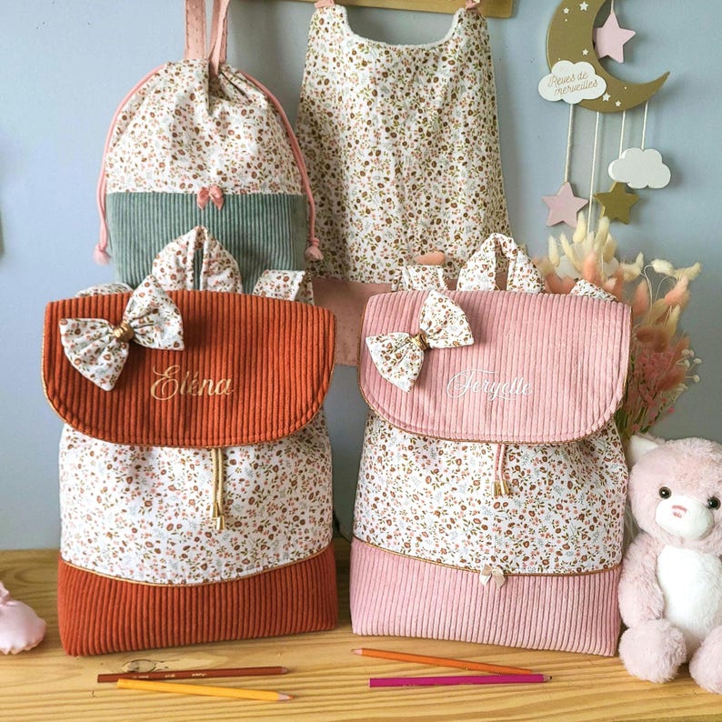 May include: Two corduroy backpacks with floral fabric panels, one in rust orange and one in light pink. Each features a matching floral bow on the flap, a drawstring closure, and embroidered names Elena and Feryelle. A matching drawstring bag and bib are visible in the background.
