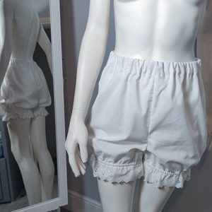 May include: White bloomers featuring an elasticized waistband and gathered leg openings finished with a delicate eyelet lace trim. The shorts have a voluminous, puffed silhouette and are displayed on a mannequin, with a mirror reflecting the back.