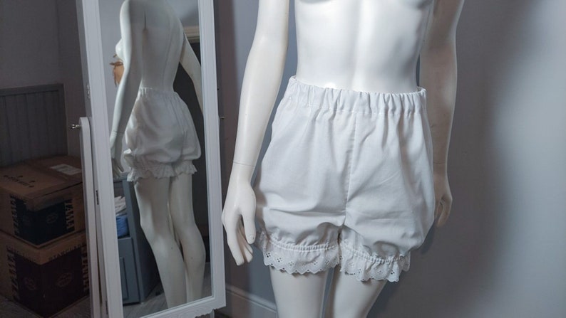 May include: White bloomers featuring an elasticized waistband and gathered leg openings finished with a delicate eyelet lace trim. The shorts have a voluminous, puffed silhouette and are displayed on a mannequin, with a mirror reflecting the back.