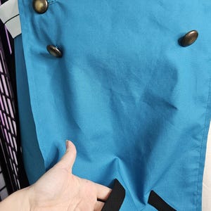 May include: A bright teal coat featuring a black collar and black trim accents. The garment is double-breasted with six round, metallic bronze-toned buttons arranged in two vertical rows. A small white tassel hangs from the lower side hem.