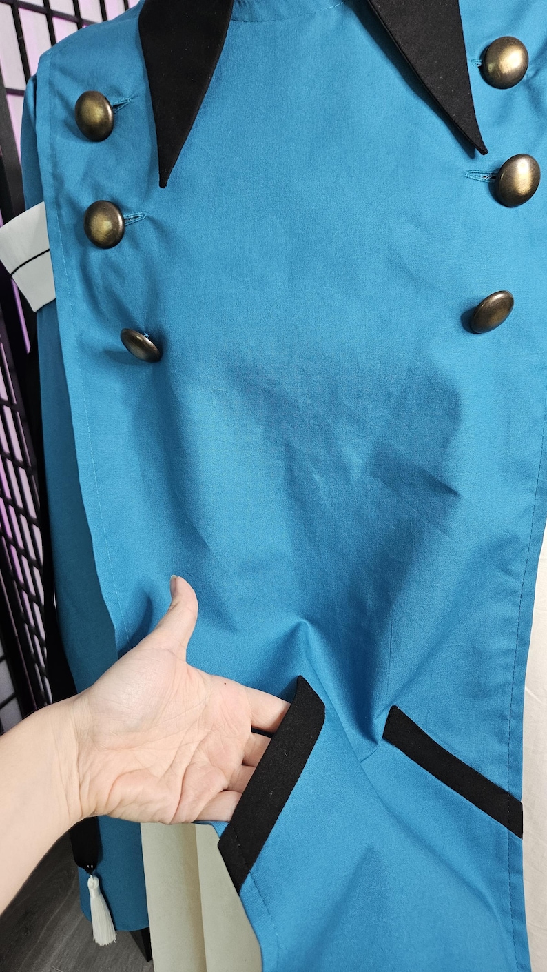 May include: A bright teal coat featuring a black collar and black trim accents. The garment is double-breasted with six round, metallic bronze-toned buttons arranged in two vertical rows. A small white tassel hangs from the lower side hem.