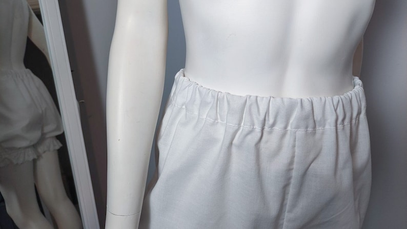 May include: A pair of plain white bloomers featuring an elasticized waistband with gathered fabric detailing. The garment is displayed on a mannequin, showing a simple, solid white design with a soft texture and a comfortable, loose-fitting silhouette.