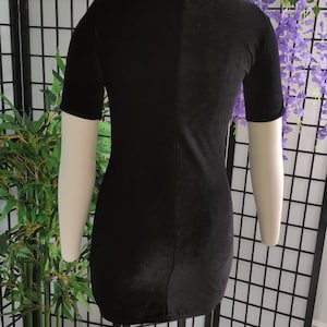 Puede incluir: Vestido negro de terciopelo con mangas cortas y cuello alto, exhibido en un maniqu&iacute;. El vestido es entallado y tiene un dise&ntilde;o sencillo. El maniqu&iacute; se encuentra frente a un biombo y plantas decorativas.