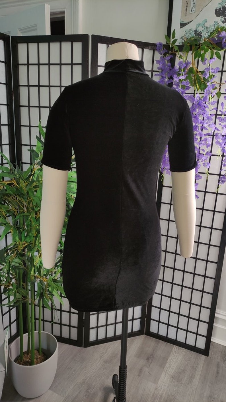 Puede incluir: Vestido negro de terciopelo con mangas cortas y cuello alto, exhibido en un maniqu&iacute;. El vestido es entallado y tiene un dise&ntilde;o sencillo. El maniqu&iacute; se encuentra frente a un biombo y plantas decorativas.