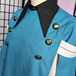 May include: A teal cape with a black collar and large brass-toned buttons. The garment features a white armband with a thin black horizontal line detail. It is displayed on a mannequin, showing a layered design with a white fabric panel underneath.