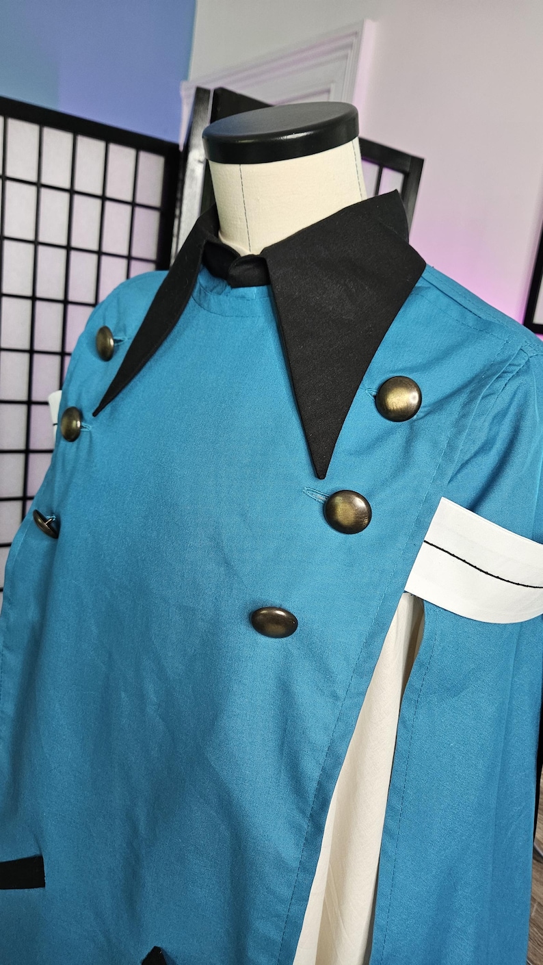 May include: A teal cape with a black collar and large brass-toned buttons. The garment features a white armband with a thin black horizontal line detail. It is displayed on a mannequin, showing a layered design with a white fabric panel underneath.