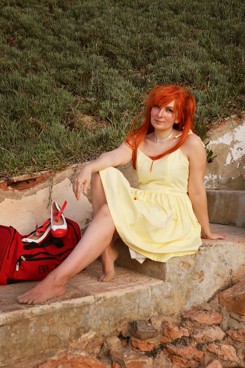 K&ouml;nnte beinhalten: Eine Frau mit langen, leuchtend orangefarbenen Haaren tr&auml;gt ein gelbes Sommerkleid und sitzt auf einer Steintreppe. Neben ihr steht eine rote Tasche mit dem Wort "EVA" darauf.