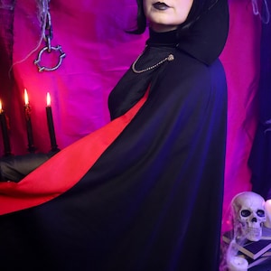May include: A black cape with a vibrant red lining, featuring a high collar and a gold-toned chain closure at the neck. The garment is worn over a black high-neck top, creating a dramatic silhouette with its contrasting interior fabric color.
