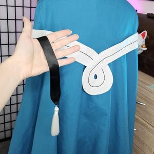May include: A teal hooded cloak featuring a white horizontal band across the back with a central circular design. A long black ribbon hangs from the white band, ending in a white tassel. The cloak is worn over a white garment, creating a layered look.