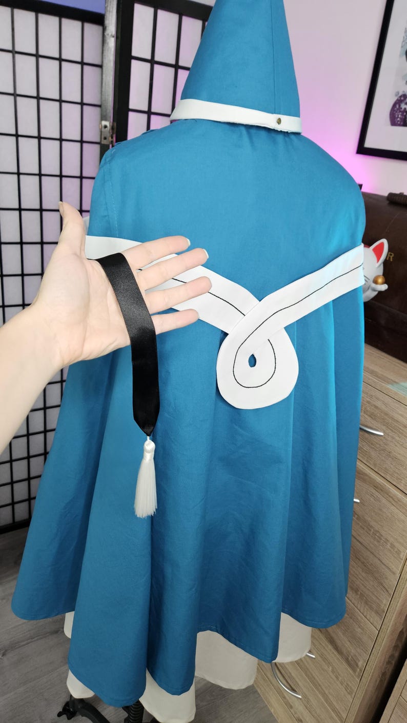 May include: A teal hooded cloak featuring a white horizontal band across the back with a central circular design. A long black ribbon hangs from the white band, ending in a white tassel. The cloak is worn over a white garment, creating a layered look.
