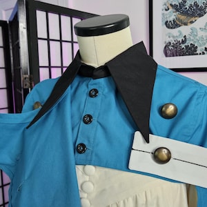 May include: A bright blue jacket with a sharp black collar and a white arm band featuring a metallic gold button. The jacket includes a front button closure with black buttons and a secondary layer of white fabric underneath the blue garment.