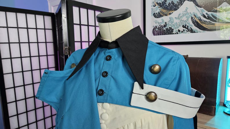 May include: A bright blue jacket with a sharp black collar and a white arm band featuring a metallic gold button. The jacket includes a front button closure with black buttons and a secondary layer of white fabric underneath the blue garment.