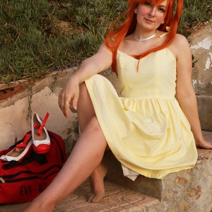 K&ouml;nnte beinhalten: Eine Frau mit langen, leuchtend orangefarbenen Haaren tr&auml;gt ein gelbes Sommerkleid und sitzt auf einer Steintreppe. Neben ihr liegt eine rote Tasche mit dem Text "EVA-02".